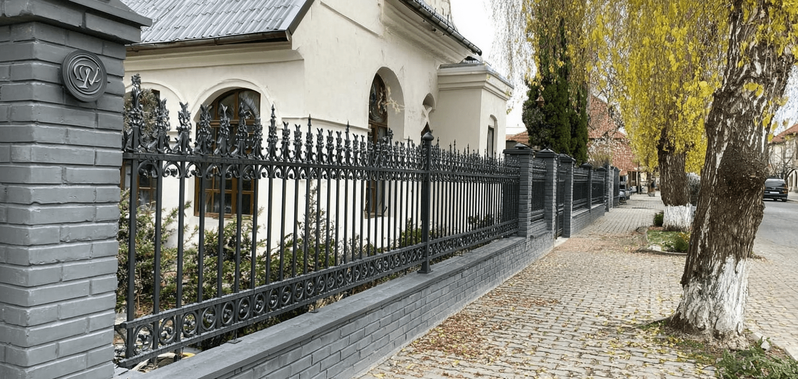 Handgeschmiedeter schwarzer Eisenzaun mit ornamentalen Verzierungen und grauen Backsteinpfeilern vor elegantem Wohnhaus